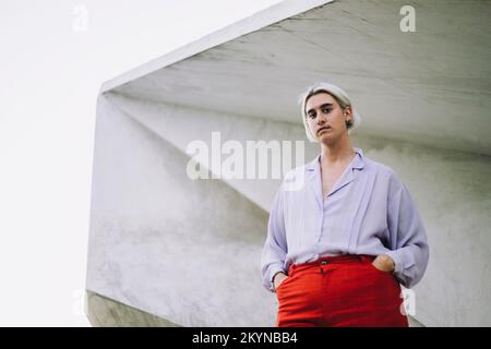 Porträt einer nicht-binären Person, die mit den Händen in Taschen an der Wand steht Stockfoto