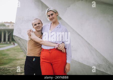 Porträt von lächelnden, nicht binären Freunden, die im Park gegen die Wand stehen Stockfoto