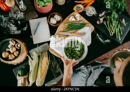 Die Hand eines weiblichen Küchenchefs hält eine Schüssel mit frischen grünen Erbsen an der Theke in der Studioküche Stockfoto