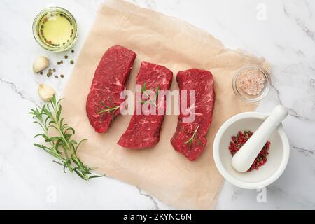 Rohe Steaks. Top Blade Steaks auf Holzbrettern mit Gewürzen, Rosmarin, Gemüse und Zutaten zum Kochen auf Pergamentpapier auf weißem Hintergrund Stockfoto