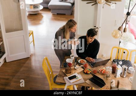 Männliche und weibliche Freiberufler, die an einem Laptop arbeiten, während sie zu Hause frühstücken Stockfoto