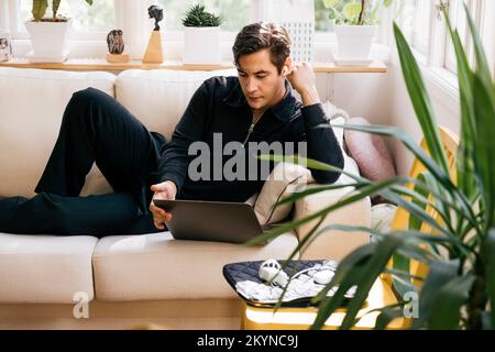 Junger männlicher Freiberufler, der zu Hause auf dem Sofa auf dem Laptop sitzt Stockfoto