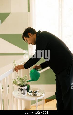 Seitenansicht eines jungen Mannes, der zu Hause Pflanzen auf einem Beistelltisch gießt Stockfoto