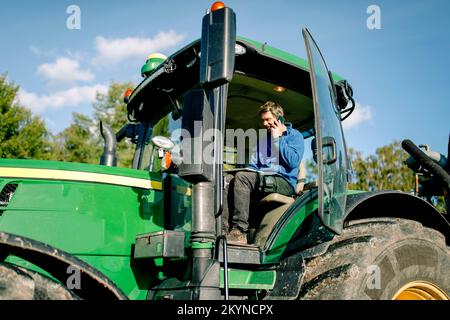Landwirt, der einen Tablet-PC verwendet und auf dem Hof im Traktor sitzt und mit dem Smartphone spricht Stockfoto