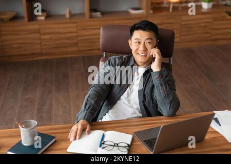 Glücklicher, erfolgreicher, erwachsener, asiatischer Geschäftsmann telefoniert, sitzt mit Computer am Tisch in der Inneneinrichtung des Heimbüros Stockfoto