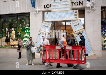 Ein Arbeiter von einer nahegelegenen Baustelle führt Sicherheitsbarrieren an den Käufern vor der Bond Street Filiale von Louis Vuitton vorbei, deren Fenster am 1.. Dezember 2022 in Mayfair, London, England, zu Weihnachten ausgestellt ist. Stockfoto