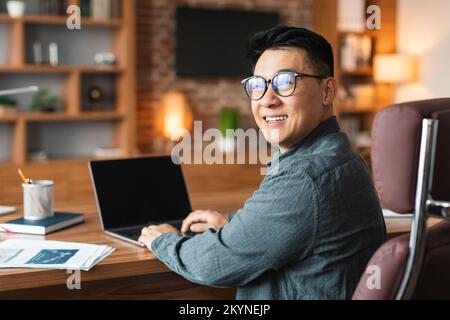 Lächelnder asiatischer Geschäftsmann mittleren Alters mit Brille arbeitet an einem Computer mit leerem Bildschirm und sitzt am Tisch im Büro Stockfoto