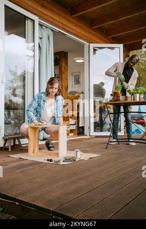 Junge Frau, die Möbel macht, mit einem Mann, der Gartenarbeit auf der Veranda macht Stockfoto