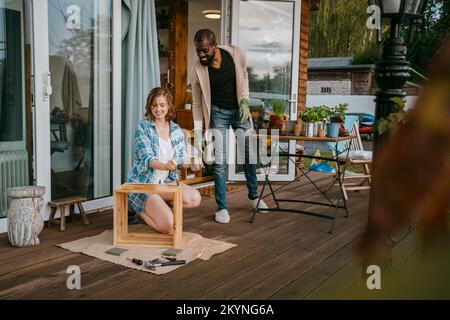 Ein erwachsener Mann, der eine Frau sieht, die Möbel auf der Veranda vor dem Haus malt Stockfoto
