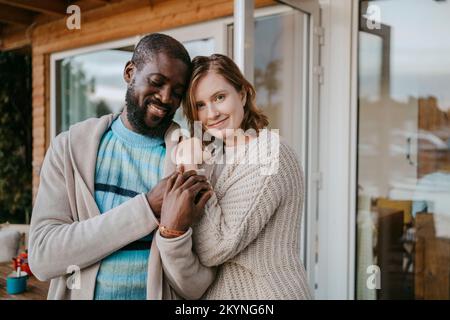 Porträt einer lächelnden jungen Frau, die den Mann umarmt, während sie auf der Veranda steht Stockfoto