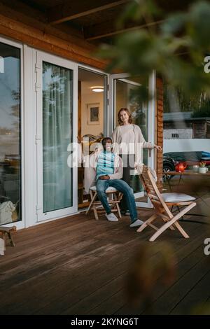 Junge Frau, die neben einem Mann steht und auf einem Stuhl auf der Veranda vor dem Haus sitzt Stockfoto