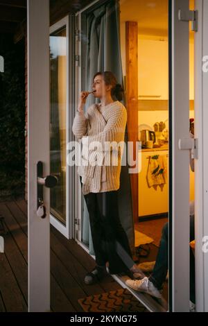 Eine junge Frau, die einen Pullover trägt und Zähne putzt, während sie an der Tür steht und durch die Glastür gesehen wird Stockfoto