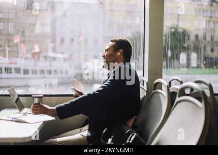 Seitenansicht eines männlichen Freiberuflers, der mit Smartphone und isoliertem Getränkebehälter am Tisch der Fähre sitzt Stockfoto