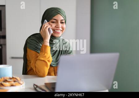 Lächelnde Mulsim-Frau, Die Zu Hause Mit Dem Laptop Telefoniert Stockfoto