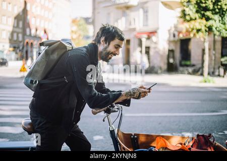 Seitenansicht eines lächelnden Geschäftsmanns, der ein Smartphone benutzt, während er sich auf ein Cargo Bike lehnt Stockfoto