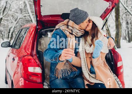 Ein verliebtes Paar küsst brennende Glitzer im Kofferraum im verschneiten Winterwald. Besucher, die Weihnachten im Freien während eines Roadtrips feiern Stockfoto