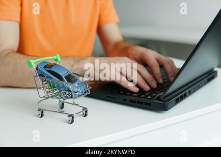 Kleiner Einkaufswagen mit Miniaturauto auf dem Tisch. Mann, der ein Notebook benutzt, um ein neues Auto online zu fahren. Stockfoto