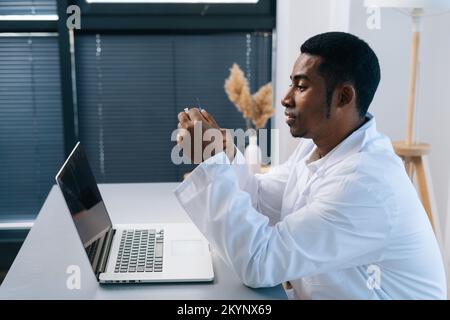 Side View sot des afroamerikanischen männlichen Arztes in weißer Uniform durchdacht zu schauen Spritze mit Impfstoff in der Hand Sitzen am Tisch mit Laptop. Stockfoto