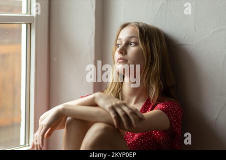 Ein romantisches Mädchen in einem roten Kleid sitzt auf dem Boden neben dem Fenster und schaut aus dem Fenster. Stockfoto