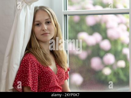 Ein junges Mädchen in einem roten Kleid sitzt auf dem Boden vor einem Busch mit rosa Blumen und schaut in die Kamera Stockfoto