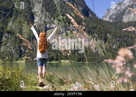 Eine schlanke, rothaarige, langhaarige Frau steht am Ufer eines klaren Sees in den Alpen Stockfoto