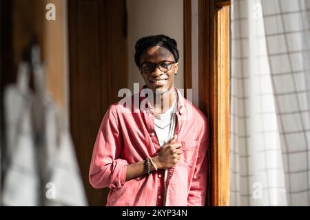 Fröhlicher Afrikaner, der positive Emotionen ausdrückt, vor der Kamera lächelt, in einem sonnigen Raum steht Stockfoto