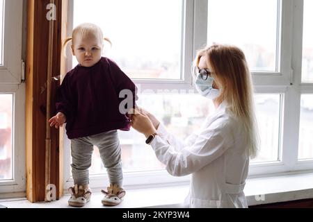 Kleines blondes Mädchen Baby steht auf dem Fensterbrett neben Kindern Arzt trägt weiße Uniform zu Hause, hält Patient Hand. Stockfoto