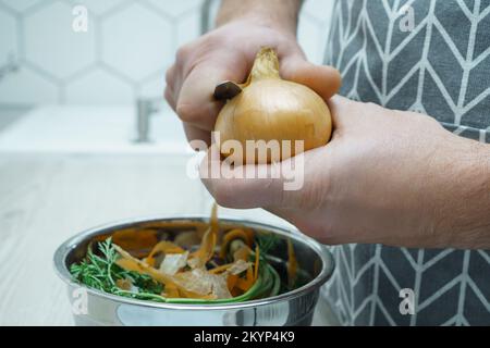 Männliche Hände schälen mit dem Messer gelbe Zwiebel über eine Schüssel Gemüseschalen. Kochen Sie auf dem Küchentisch, bereiten Sie Zutaten zu, verschrotten Sie das Essen. Gesunder Irrer Stockfoto