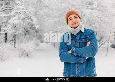 Porträt eines stilvollen Mannes im verschneiten Winterpark mit Jeans Mütze, der draußen posiert. Gut aussehende Blicke in die Kamera. Männermode. Leerzeichen Stockfoto