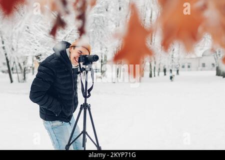 Fotograf, der Fotos mit einer professionellen Digitalkamera auf einem Stativ im verschneiten Winterpark macht und in den Sucher schaut. Landschaftsbilder. Outdoor-Fotografie Stockfoto