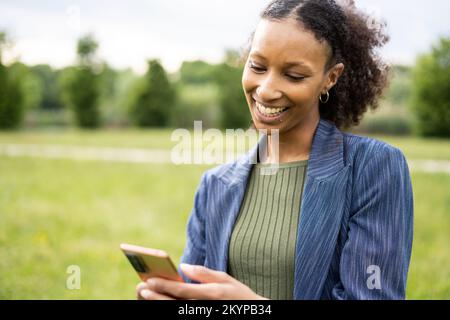 Eine junge karibikfrau liest eine Nachricht seines besten Freundes und lächelt, während sie einen schönen Tag im Freien genießt. Stockfoto