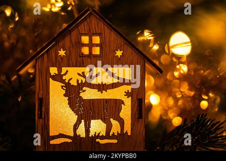 Weihnachtsdekoration in Form eines Hauses mit einem Rentier im Inneren, beleuchtet durch das warme Licht elektrischer Girlanden auf einer Tanne. Schöner, verschwommener Hintergrund. Stockfoto