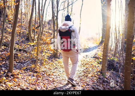 Sportlicher Vater, der seinen kleinen Sohn mit Winteranzug und Mütze im Rucksackträger beim Wandern im Herbstwald trug Stockfoto