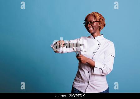 afroamerikanische Chefkochin in Uniform schärft professionelle Messerklinge, bevor sie ihre Schicht beginnt. Frau, die Küchenutensilien für das Kochen zubereitet, scharfe Küchengegenstände in den Händen hält. Stockfoto