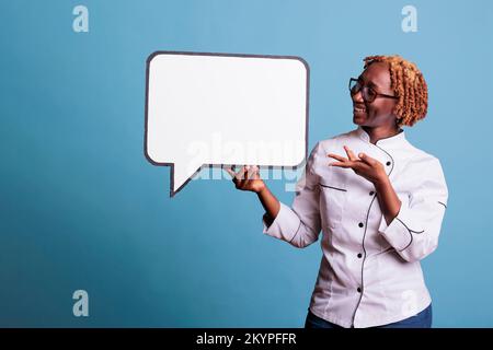 Lächelnde Küchenangestellte, die auf eine leere dialogwolke mit Kopierraum zeigt. Koch hält dialogblase mit Nachrichtenrahmen, der auf die Kamera schaut, Studioaufnahme auf blauem Hintergrund. Stockfoto