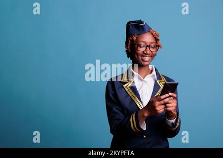 Lächelnde weibliche Flugbegleiterin in Uniform, die Nachrichten auf dem Handy liest. Unterhaltsame Stewardess mit Handheld-Gerät im Studio vor blauem Hintergrund. Stockfoto