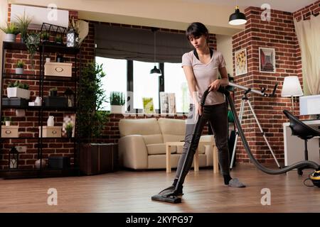Die Ehefrau kümmert sich um den Frühjahrsputz, saugt den Staub im Wohnzimmer zu Hause ab. Hausfrau in Hausschuhen macht Hausarbeiten mit Hingabe, räumt auf und putzt alles. Stockfoto