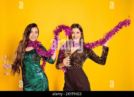 Zwei wunderschöne junge Frauen in Cocktailkleidern mit Champagnergläsern in den Händen, die lächeln und rosa Boas schwenken Stockfoto