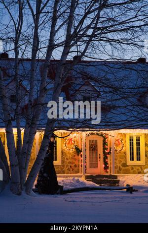 Beleuchtetes zweistöckiges Haus aus Feldstein im Canadiana-Stil dekoriert mit Weihnachtskränzen und Lichtern in der Dämmerung. Stockfoto