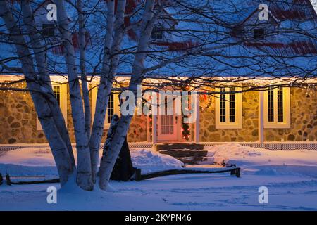 Beleuchtetes zweistöckiges Haus aus Feldstein im Canadiana-Stil dekoriert mit Weihnachtskränzen und Lichtern in der Dämmerung. Stockfoto