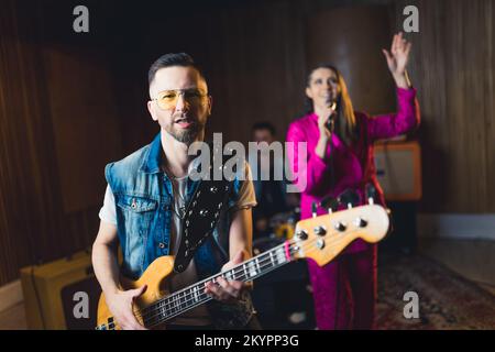 Ein mittelgroßer bärtiger Mann in einer gelben Glasbrille spielt auf einer E-Gitarre. Unscharfer weiblicher Sänger und männlicher Schlagzeuger im Hintergrund. Konzert-Konzept im Innenbereich. Hochwertiges Foto Stockfoto