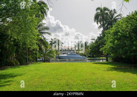 Blick auf eine Yacht mit Abdeckung von einem Hof mit grünem Rasen und Zaun an der Bucht in Miami, Florida. Blick von einem Rasen mit Bäumen an der Seite über einem großen Stockfoto