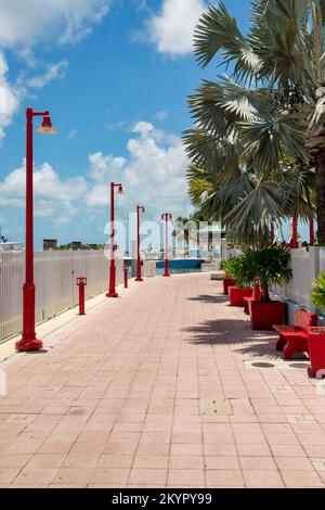 Bürgersteig an der Bucht mit zeitgenössischen roten Straßenlaternen, Bänken und Pflanzmaschinen in Miami, FL. Es gibt einen Zaun in der Nähe des Wassers mit Booten an den Docks A. Stockfoto