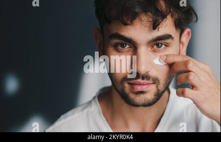 Kopfschuss eines gutaussehenden jungen Mannes, der Anti-Aging-Feuchtigkeitscreme auf Gesicht aufträgt, isoliert im Studiohintergrund und in die Kamera schaut. Konzept der Fairness und Stockfoto