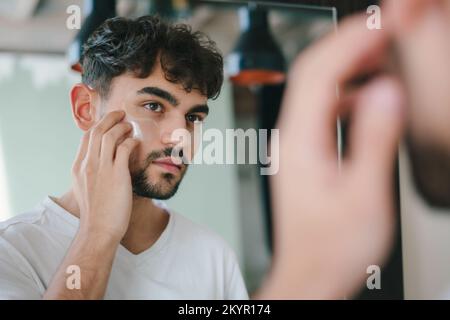 Ein bärtiger Mann, der Augenflecken unter die Augen legt, während er in den Spiegel schaut. Falten und Gesichtspflege für Männer. Hautpflegeroutine. Home Spa Care-Konzept Stockfoto