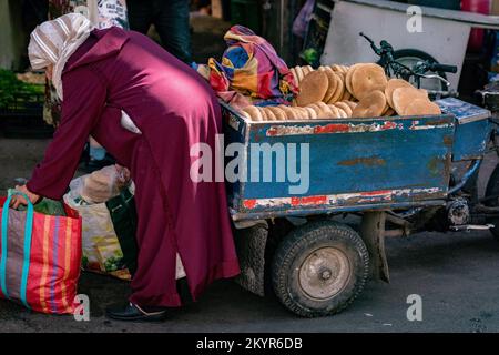 Eine marokkanische Frau kauft frisch gebackenes Khobz (marokkanisches Fladenbrot in Scheibenform) von einem Motorradverkäufer im Souk in der Medina von Marrakesch, Marokko Stockfoto