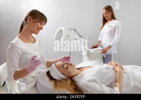 Kosmetologe, der eine Maske aus Lehm auf das Gesicht einer schönen Frau anwendet. Spa-Behandlung und Gesichtspflege im Schönheitssalon Stockfoto