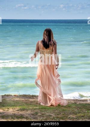 Eine junge Frau in einem fließenden formellen Kleid steht auf einem Hügel hoch über einem aktiven See und sieht aus, als würde sie springen Stockfoto