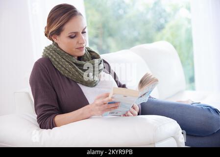 In einen guten Roman vertieft. Eine junge Frau, die auf ihrer Couch sitzt und ein Buch liest. Stockfoto