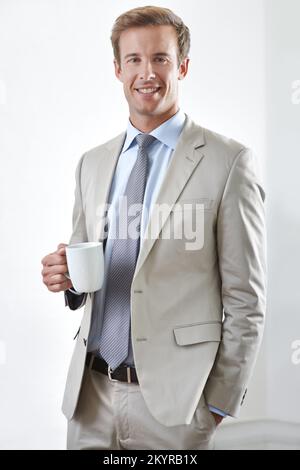 Ich genieße eine Kaffeepause. Ein junger Geschäftsmann, der mit einer Tasse Kaffee in der Hand steht und in die Kamera lächelt. Stockfoto